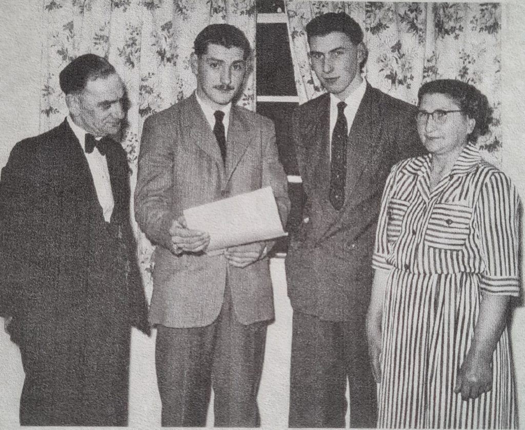 Black and white picture of four people, a middle-aged man on left, two young men in centre, and middle-aged woman on right, in 1950s suits. The young man in centre holds a piece of paper. They look proud.